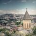 View of a historical church tower with Tbilisi's cityscape in the background under a blue sky.