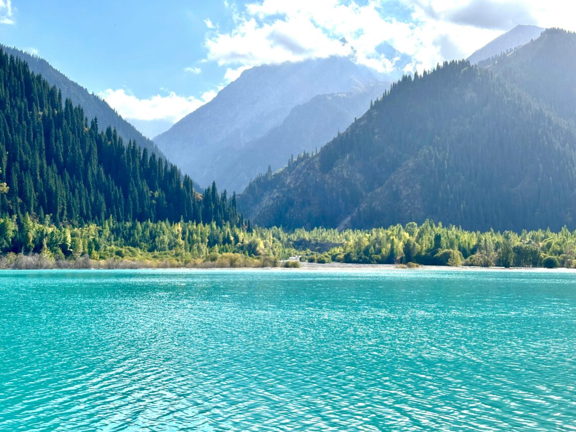 Beautiful turquoise lake surrounded by mountains and forests in Kazakhstan, under a clear blue sky.